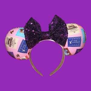 May include: A headband featuring two round ears covered in a light pink fabric printed with various rectangular labels including Liquorice Wands and Chocolate Frog. A large, central bow made of dark purple sequins sits between the two ears.