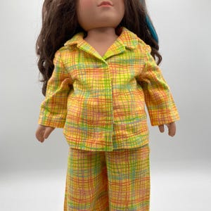 May include: A doll wearing a yellow, orange, and green plaid pajama set. The doll has brown hair with blue highlights and blue eyes. The pajama set includes a collared shirt and matching pants.