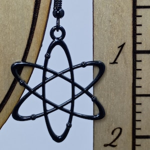 May include: Black atom-shaped earring with a black ear wire. The earring is a stylized representation of an atom, with a central oval and intersecting lines. The earring is approximately 1 inch in length.