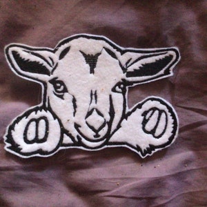 May include: A white and black embroidered patch featuring a cartoon goat's head. The goat has large ears and a detailed face, with black outlines and accents. The patch is likely for clothing or accessories.