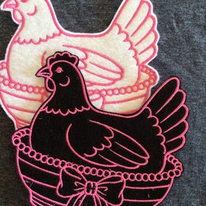 May include: Two felt chicken appliqués. One is white with pink trim, the other is black with pink trim. Both are in decorative baskets with pink beaded trim and a pink bow. The background is a solid gray.