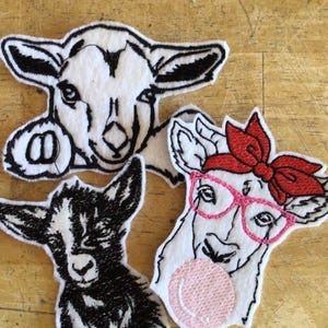 May include: Three goat-shaped felt appliques. One is black and white, another has a red bow and pink glasses, and the third is a black and white outline. The appliques are on a wooden surface.
