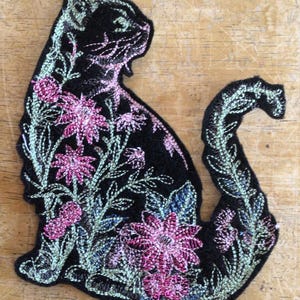 May include: A black cat-shaped embroidered patch featuring intricate floral designs in shades of pink and light green. The embroidery covers the body and tail of the cat, showcasing detailed petals, leaves, and stems stitched onto a black fabric base.