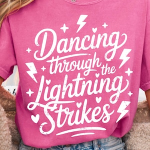 Puede incluir: Una camiseta rosa con el texto blanco "Dancing through the Lightning Strikes". El diseño incluye rayos, estrellas y corazones. La camiseta es de manga corta y cuello redondo.