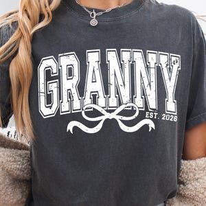 May include: Dark gray t-shirt with the word "GRANNY" in white, collegiate-style lettering. Below the word is a white bow design, and the text "EST. 2026". The shirt is a casual style.