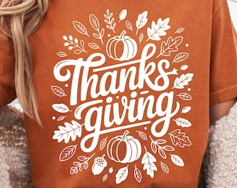 May include: Rust-colored t-shirt with the text "Thanksgiving" in white script, surrounded by leaf and pumpkin illustrations. A fall-themed graphic tee for the holiday season.