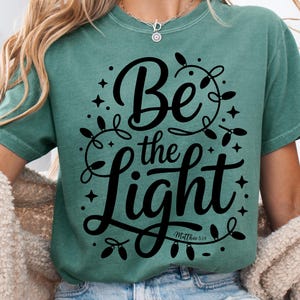 May include: A teal-colored t-shirt with the words "Be the Light" in a black script font, surrounded by decorative string lights and stars. The text "Matthew 5:14" is below the main text. The shirt has short sleeves.