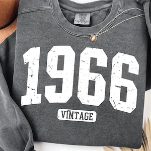 Könnte beinhalten: Dunkelgraues Sweatshirt mit der Jahreszahl "1966" in großen, weißen Buchstaben im Used-Look. Darunter steht das Wort "VINTAGE". Eine goldene Halskette und ein brauner Hut sind zu sehen. Das Sweatshirt ist von der Marke Comfort Colors.