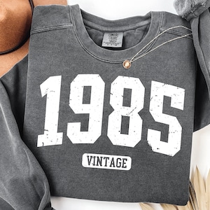 May include: Dark gray sweatshirt with the year "1985" in large white block letters and the word "VINTAGE" underneath. A gold pendant necklace rests on the sweatshirt. The sweatshirt is folded on a white surface.