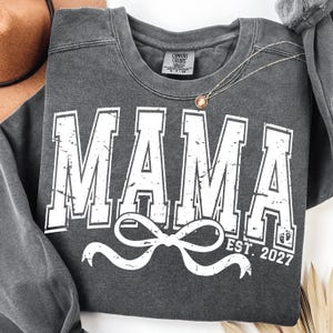 Peut inclure: Un sweat-shirt gris anthracite avec le mot "MAMA" en lettres blanches et usées. En dessous, un motif de nœud blanc et le texte "EST. 2027" avec une empreinte de pied de bébé. Un collier pendentif doré est posé sur le sweat-shirt.