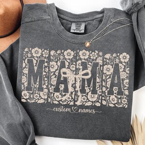 Puede incluir: Sudadera gris oscuro con la palabra "MAMA" en letras grandes, adornada con diseños florales. Un lazo está centrado en medio de la palabra. Debajo de la palabra "MAMA" está el texto "custom names" en cursiva.