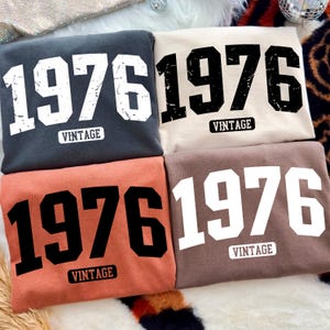 May include: Four folded sweatshirts in various colors: charcoal, beige, coral, and brown. Each shirt features the number "1976" in a bold font and the word "VINTAGE" underneath.