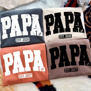 May include: Four folded sweatshirts with the word "PAPA" in large, distressed lettering. Each shirt has "EST. 2026" or "EST. 2027" printed below. The shirts are in shades of gray, cream, coral, and brown, with a small footprint graphic.