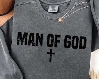 Op de afbeelding: Een antracietgrijs sweatshirt met de tekst "MAN OF GOD" in vette, zwarte letters en een zwart kruis eronder. Een goudkleurige ketting met een hanger ligt op het shirt. Het shirt is gemaakt van een zacht materiaal.