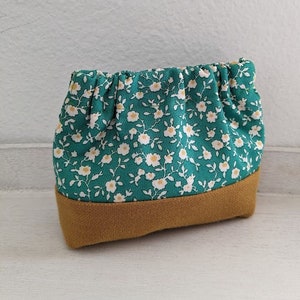 Könnte beinhalten: Eine kleine grüne Tasche mit einem weißen Blumenmuster und einem braunen Boden. Die Tasche ist aus Stoff und hat einen gerafften oberen Rand.
