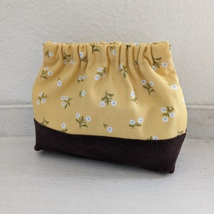 Könnte beinhalten: Eine kleine gelbe Stofftasche mit einem weißen Blumenmuster und einem braunen Boden. Die Tasche hat einen gerafften oberen Rand und ist aus einem weichen, baumwollartigen Material gefertigt.