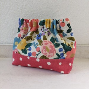 Könnte beinhalten: Eine kleine, rosa-weiße Stofftasche mit floralem Muster und einem weißen Punktmuster am Rand. Die Tasche ist oben gerafft und hat einen Kordelverschluss.