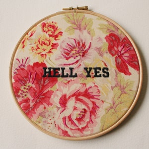 May include: A round embroidery hoop with a floral fabric background. The fabric has a pink, red, and yellow floral pattern. The words "HELL YES" are embroidered in black thread on the fabric.