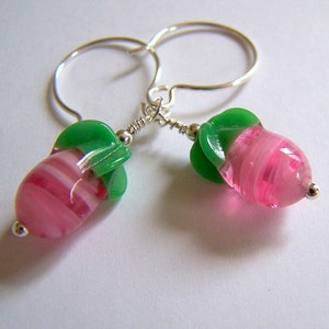 May include: A pair of handmade earrings featuring pink and white striped glass beads, topped with green glass accents. The earrings are suspended from silver-colored ear wires and have small silver-colored beads at the bottom.