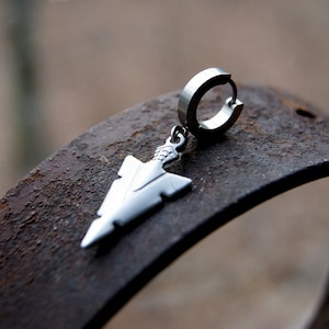 May include: Silver hoop earring with a silver arrowhead pendant. The earring is on a rusty metal surface.