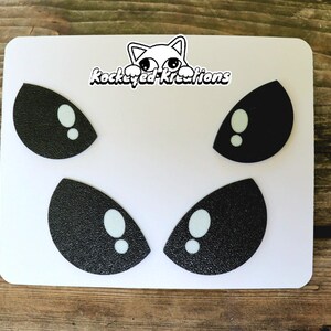 Puede incluir: Una pegatina rectangular blanca con cuatro pares de ojos de dibujos animados en blanco y negro. Los ojos tienen una textura brillante y est&aacute;n dispuestos en dos filas. La pegatina tambi&eacute;n incluye el texto "Rockeyed-Kreations" y un dibujo de gato.