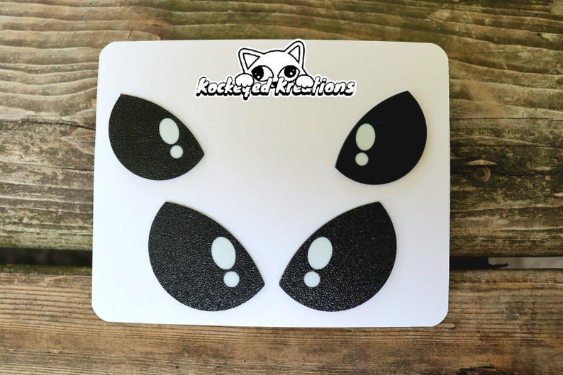 Puede incluir: Una pegatina rectangular blanca con cuatro pares de ojos de dibujos animados en blanco y negro. Los ojos tienen una textura brillante y est&aacute;n dispuestos en dos filas. La pegatina tambi&eacute;n incluye el texto "Rockeyed-Kreations" y un dibujo de gato.