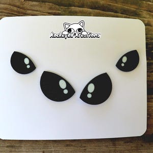 Puede incluir: Una tarjeta blanca con formas de ojos negros y reflejos blancos. La tarjeta tiene esquinas redondeadas y un logotipo "kockeyed kreations" con una ilustraci&oacute;n de gato.