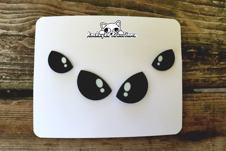 Puede incluir: Una tarjeta blanca con formas de ojos negros y reflejos blancos. La tarjeta tiene esquinas redondeadas y un logotipo "kockeyed kreations" con una ilustraci&oacute;n de gato.