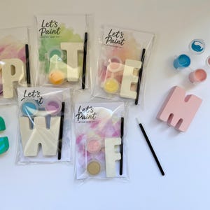 May include: A collection of letter paint kits and individual letters. The kits include paint, brushes, and wooden letters. The letters are white, pink, and green. The kits are packaged in clear plastic bags with the text "Let's Paint".