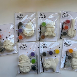 May include: Six individual Let's Paint Halloween paint kits in clear plastic packaging. Each kit includes a white ceramic figure, three small paint pots in various colors, and a thin black paintbrush on a card with orange and black confetti.