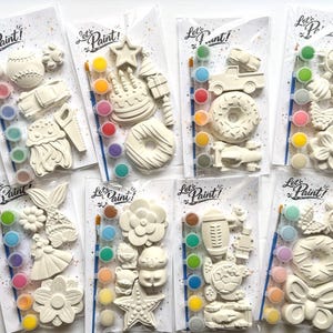 May include: Eight individual Let's Paint! craft kits in clear plastic packaging. Each kit contains white plaster figures in various shapes like donuts, flowers, animals, and vehicles, along with a small paintbrush and a strip of six paint pots.