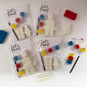 May include: Paint kits with the text "Let's Paint" on the packaging. Each kit includes a white Lego-style figure, paint pots in blue, red, and yellow, a paintbrush, and a Lego brick. Additional Lego bricks and figures are scattered around.