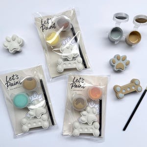 May include: Dog paint kits with paw print and bone-shaped molds, paint pots, and paintbrushes. The kits are packaged in clear plastic bags with the text "Let's Paint". The paint colors include gray, yellow, brown, and orange.