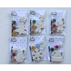 May include: Six Let's Paint Christmas paint kits in individual clear plastic packaging. Each kit contains a white ceramic ornament, a small paintbrush, and three small paint pots in various colors. The ornaments include a tree, gingerbread person, snowman, snowflake, and Santa hat.