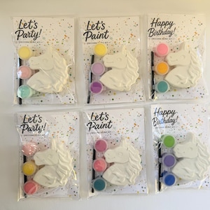 May include: Six individual unicorn paint kits packaged in clear plastic. Each kit includes a white ceramic unicorn head, a small black paintbrush, and three small circular paint pots in various colors. The backing cards feature confetti patterns and text reading Let's Party, Let's Paint, or Happy Birthday.