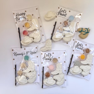 May include: Five clear plastic bags containing horse paint kits, each with white ceramic figures of cowboy boots, hats, and horses, along with small paint pots and a black brush. The packaging features text including Happy Birthday, Let's Paint, and Kennedy's 8th Rodeo.
