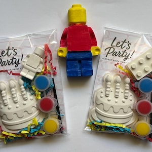 May include: Two clear plastic bags containing white plaster figures of a birthday cake and a small robot, three small paint pots in blue, red, and yellow, and colorful paper shreds. A painted yellow, red, and blue plastic figure stands above.