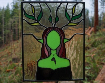 May include: A stained glass window depicting a green-skinned woman with long red hair and a black dress. The woman is surrounded by green leaves and branches. The window is framed in black metal and hangs from a chain.
