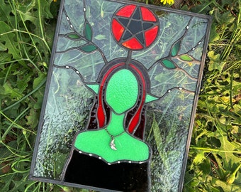 May include: A stained glass window depicting a green-skinned woman with red hair and a crescent moon necklace. The woman is surrounded by green leaves and a red and black pentagram.