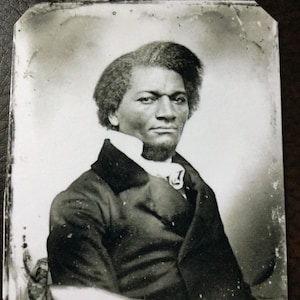 May include: Black and white portrait photograph of a man wearing a dark suit and a white collar. He has short, curly hair and a serious expression.
