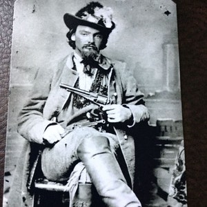 May include: A black and white photograph of a man wearing a hat and holding a gun. He is sitting in a chair with his legs crossed. The photo is in a vintage style.