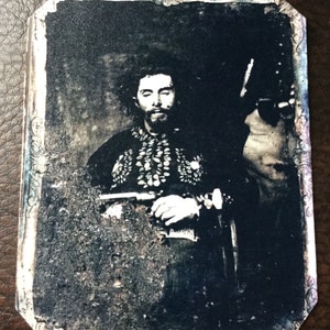May include: A black and white vintage photograph of a man wearing a dark jacket with a beaded pattern. He is holding a gun in his right hand. The photo is framed with a decorative border.