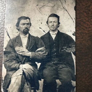 May include: Black and white photograph of two men sitting side by side. The man on the left has a beard and is wearing a dark jacket and pants. The man on the right is wearing a dark suit and has a serious expression.