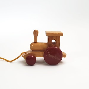 May include: A light brown wooden pull toy train with a smooth finish. The train features two dark red wheels, with the larger front wheel displaying the text Neiman Marcus in cursive script. A yellow string is attached to the front of the engine.