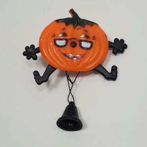 May include: A plastic orange pumpkin ornament with a green stem, black limbs, and a smiling face wearing white-rimmed glasses. The pumpkin has two black arms and two black legs extending outward, and a black bell hangs from the bottom by a string.