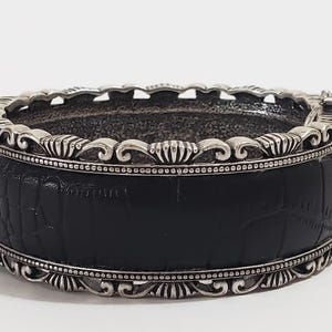 May include: A wide, black bracelet with a textured, crocodile-like pattern. The bracelet is framed with ornate silver-toned metal detailing. The design features a series of repeating patterns and decorative elements.