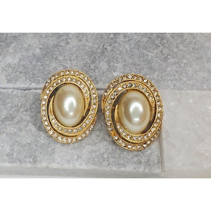 Puede incluir: Un par de pendientes de clip de tono dorado con un centro de perla falsa y un círculo de piedras de cristal transparentes alrededor de la perla.