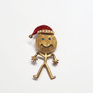 May include: A gold-coloured stick figure brooch with a round head and a smiling face. The head features blue gem eyes and a red Santa hat trimmed with faux pearls. The brooch has outstretched arms and legs.
