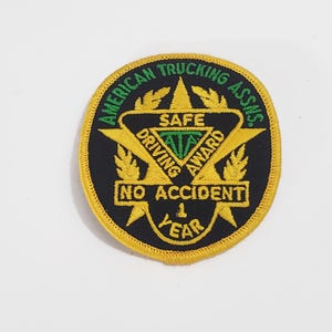 May include: A circular patch with the text "AMERICAN TRUCKING ASSNS." in green, arched around the top. The center features a gold star with "SAFE DRIVING AWARD" and "NO ACCIDENT 1 YEAR" in white and green. The patch has a black background and a gold border.
