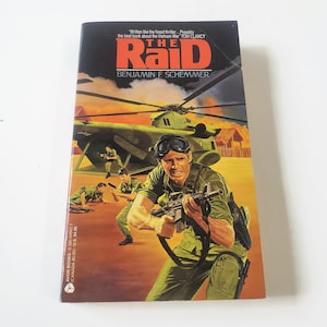 Pode incluir: Um livro de bolso intitulado The Raid de Benjamin F. Schemmer. A capa apresenta um helicóptero militar verde, soldados em uniformes verdes segurando rifles e uma paisagem desértica. O título está em letras grandes de cor laranja.